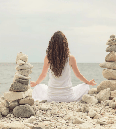 A woman sitting on a rocky beach meditating, beside here are tock towers stacked up 