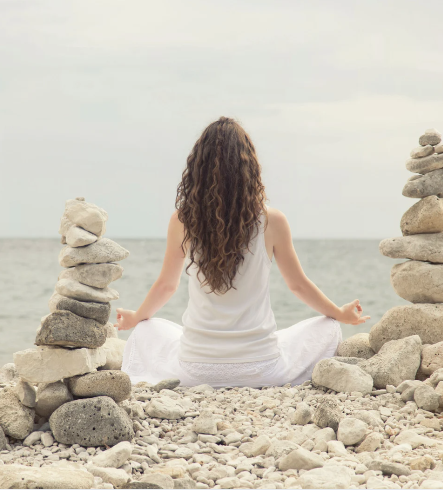 A woman sitting on a rocky beach meditating, beside here are tock towers stacked up 